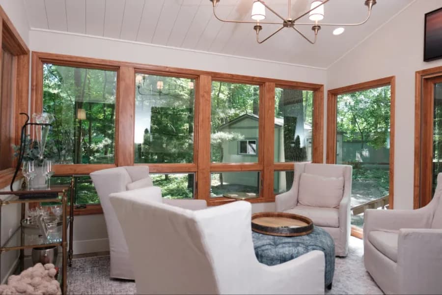 Sunroom addition with wood-framed windows overlooking a wooded yard, white shiplap ceiling, a brass chandelier and slipcovered armchairs