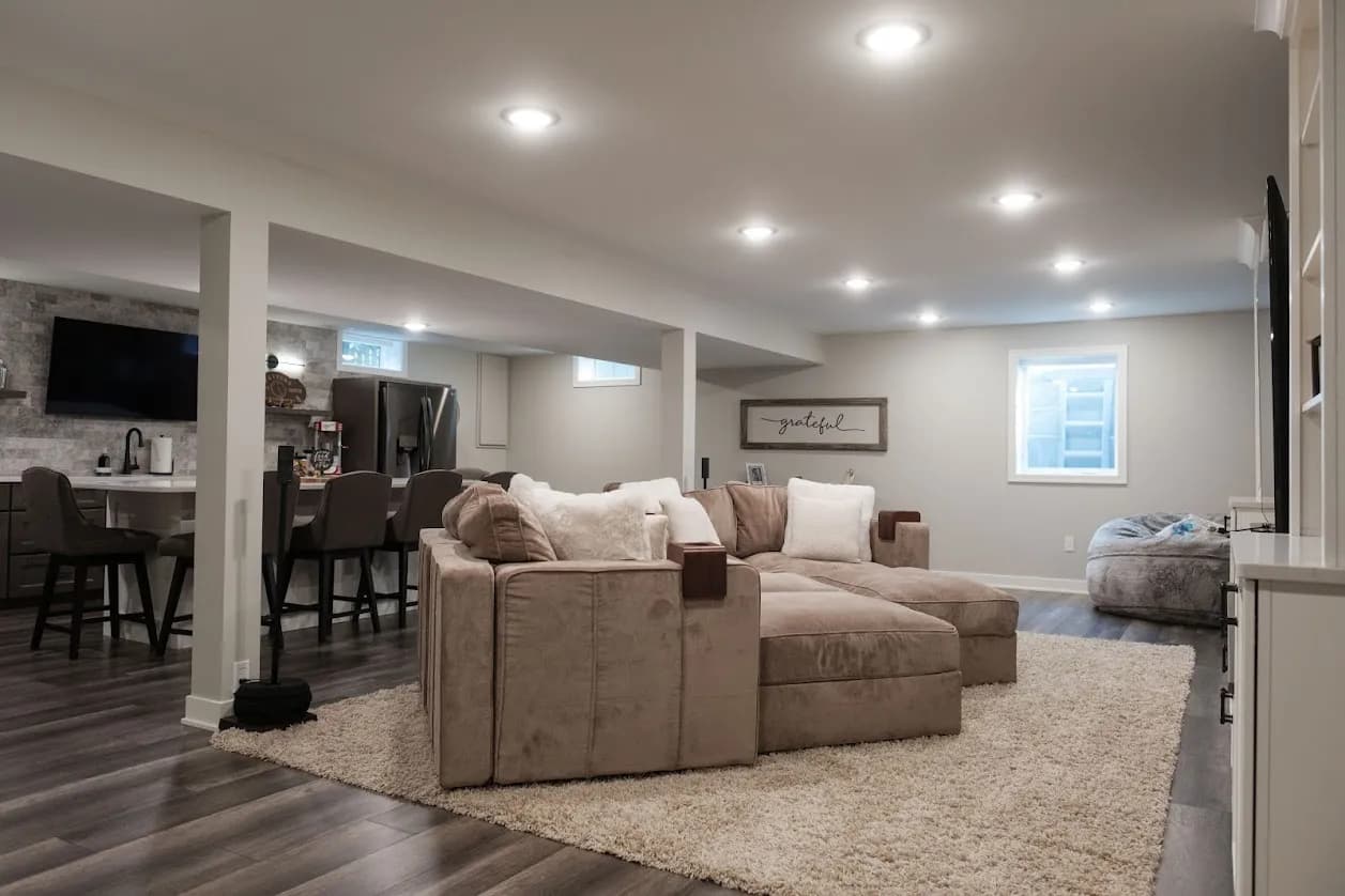 Spacious finished basement living area with a sectional sofa, wet bar and wall-mounted TV, warmly lit with recessed lighting
