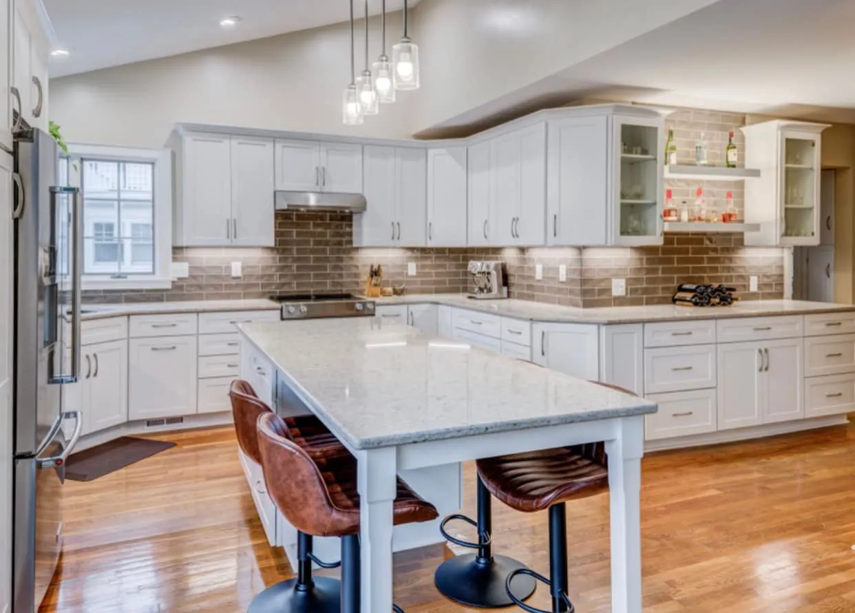 Open-concept kitchen remodel with white shaker cabinetry, a light granite island with leather counter stools, grey subway-tile backsplash and hardwood floors